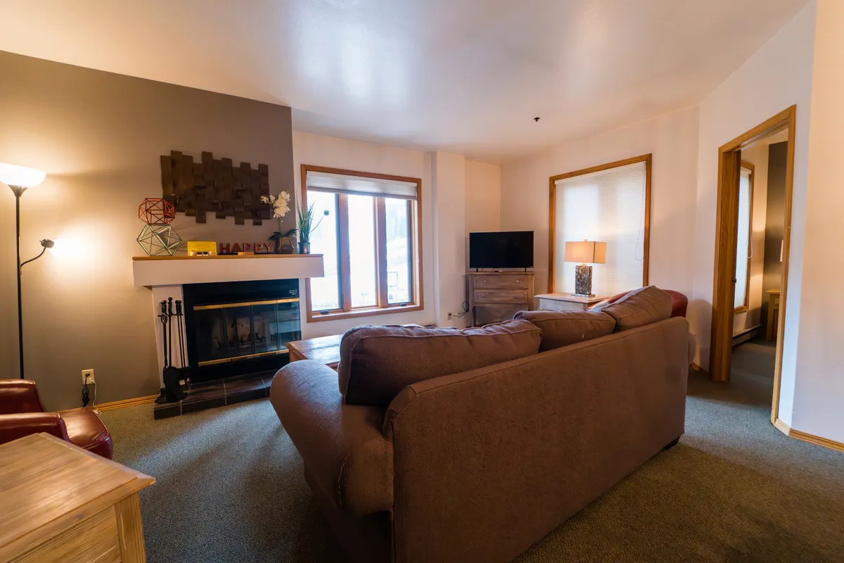 A cozy living area featuring a plush sofa set beside a wood-burning fireplace. A TV and lamp provide additional comfort, while large windows let in natural light and offer glimpses of the outdoors. The arrangement invites guests to relax, watch TV, read, or enjoy casual conversation by the fire.