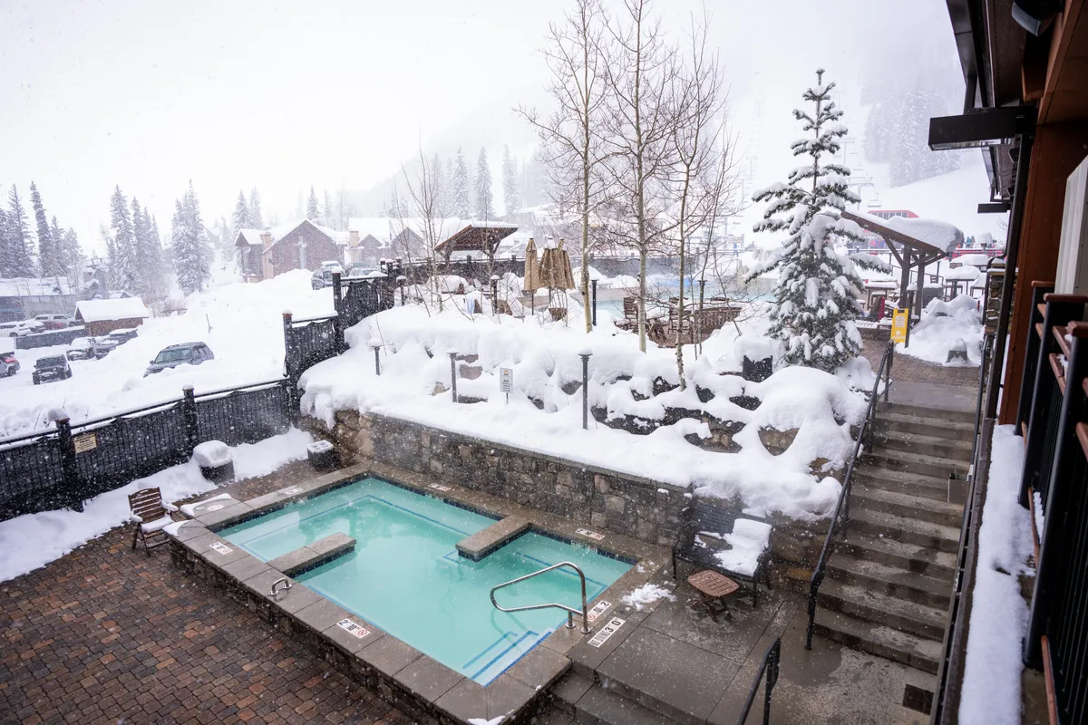 A tranquil outdoor hot tub set against a snow-covered landscape with pine trees. Steps lead down to the inviting waters, while steam rises and snow gently falls. The setting feels private, peaceful, and the perfect place for après-ski relaxation or stargazing with family.