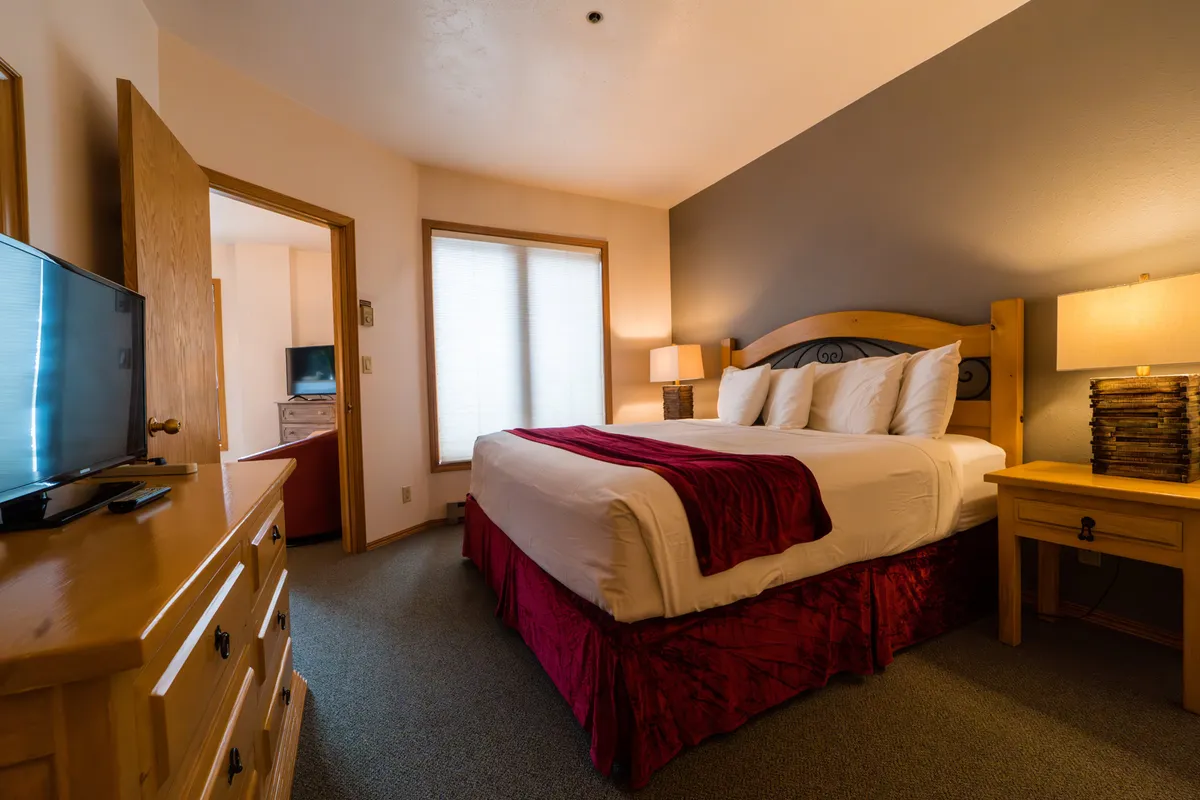 A cozy bedroom featuring a bed topped with a red throw, a side table with a lamp, and a TV on the dresser. The soft lighting and comfortable furnishings invite relaxation and restful sleep.