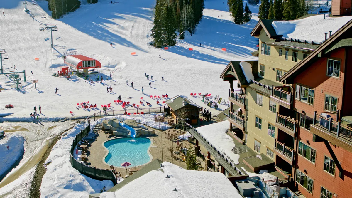 A lively exterior view showcasing skiers on the slopes, a bustling resort building, the ski lift in operation, and a heated swimming pool where guests relax, even amid the snow. The scene buzzes with energy, capturing both winter sports excitement and the unique amenity of an outdoor pool.