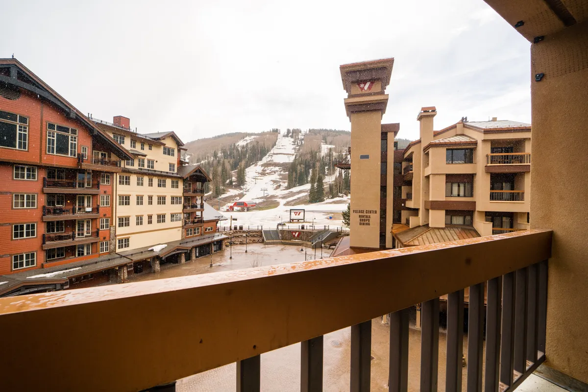 A cozy mountain village scene with ski slopes just beyond the resort buildings, groups of skiers and snowboarders in action, and balconies overlooking the adventure. The surrounding mountains create a dramatic backdrop, while the village center buzzes with activity and warmth.
